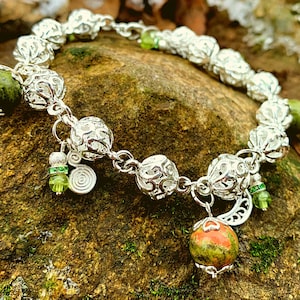 Puede incluir: Un brazalete de plata con abalorios de piedras preciosas verdes y naranjas. El brazalete presenta intrincados diseños de filigrana y abalorios colgantes.