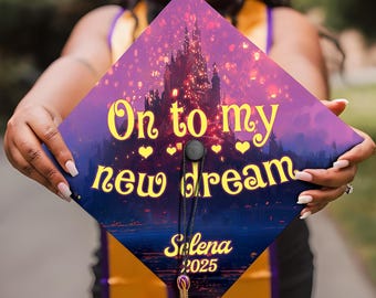Op de afbeelding: Een paarse en blauwe afstudeerhoed met een afbeelding van een kasteel en lantaarns. De tekst "On to my new dream" is in geel met hartjes geschreven. De naam "Selena" en het jaar "2025" staan ​​onderaan in geel.