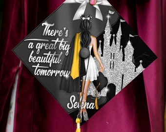 May include: A black graduation cap with a silver glitter castle design and a black and yellow illustration of a woman with a mouse ear headband. The text "There's a great big beautiful tomorrow" is written on the cap. The name "Selena" is written on the bottom of the cap.