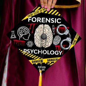 May include: Black graduation cap with yellow and black striped tape around the edge. The cap has a graphic of a brain, handcuffs, a fingerprint, and a test tube. The text "Forensic Psychology" is printed in white letters on the cap. The text "Danger" is printed in yellow letters on the cap. The text "David 2-25" is printed in white letters on the cap.