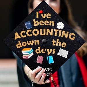 May include: Black graduation cap with gold glitter and the text "I've been ACCOUNTING down the days". The cap has a red apple, a clock, and a stack of books on it.