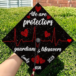 May include: Black graduation cap with a red heartbeat graphic and the text "We are protectors guardians & lifesavers" in white. The cap also has a red heart, the name "Emily" and the year "2025" in white. The cap has a red tassel with the letters "BSN" in white.