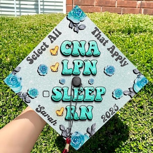 May include: A graduation cap with a white background and blue floral designs. The text "Select All That Apply" is written on the cap, along with the words "CNA", "LPN", "SLEEP", and "RN" in a turquoise color. The text "Sarah 2023" is also written on the cap.