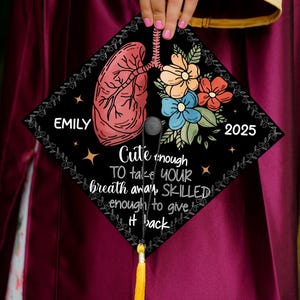 May include: Black graduation cap with a floral design and a drawing of a lung. The text on the cap reads "Cute enough to take your breath away, Skilled enough to give it back." The name "Emily" and the year "2025" are also on the cap.