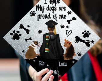 May include: A silver graduation cap with a glitter finish. The cap features a black silhouette of a person wearing a graduation cap and gown, with two dogs sitting on either side. The text "I Hope My Dogs Are Proud Of Me" is written on the cap, along with the names "Rio" and "Lulu".