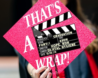 May include: Pink graduation cap with a black and white film clapboard design. The clapboard says "That's a Wrap!" and has the text "Graduation" with the details "Production, Scene, Director, Camera, Date" and the text "Take, ASDH, Hope, RDH, Dental Hygiene" with the date "11.10.24".