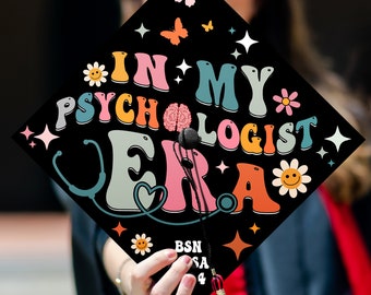 May include: A black graduation cap with the text "In My Psychologist Era" in colourful letters. The cap has a stethoscope design and a small brain graphic. The text "BSN '24" is also visible on the cap.