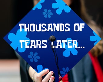 May include: A graduation cap with the text "THOUSANDS OF TEARS LATER..." in white. The cap is dark blue with light blue flower designs. The tassel is black with red and gold accents.