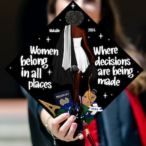 May include: Black graduation cap with the text "Women belong in all places" and "Where decisions are being made". The cap features a black and white illustration of a woman in a white dress and a black cape, holding a passport and a globe. The cap also has a pink star design.