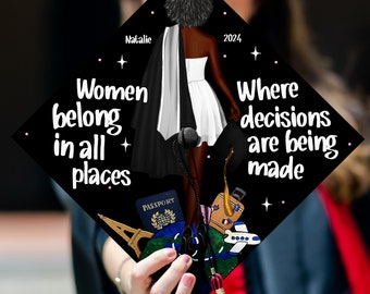 May include: Black graduation cap with the text "Women belong in all places" and "Where decisions are being made". The cap features a black and white illustration of a woman in a white dress and a black cape, holding a passport and a globe. The cap also has a pink star design.