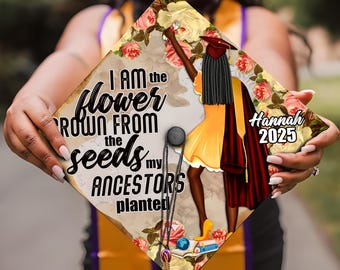 May include: A graduation cap with a floral design and the text "I am the flower grown from the seeds my ancestors planted". The cap also features a black woman in a yellow dress and a red graduation gown. The name "Hannah 2025" is printed on the cap.