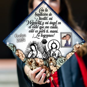 May include: A white graduation cap with blue trim and a floral design. The cap has a picture of the Virgin Mary and Jesus Christ, as well as a small picture of an older woman. The text on the cap reads "Con la bendición de Diosito, mi Virgencita y mi ángel en el cielo que me cuida, esto es para ti, mami. ¡Lo logramos!" and "Evelyn 2024".
