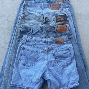 Puede incluir: Una pila de vaqueros y pantalones cortos de mezclilla azul claro. Los vaqueros tienen el clásico diseño de cinco bolsillos con parches de cuero marrón y el logotipo de Levi Strauss & Co. Los pantalones cortos también son de mezclilla azul claro.