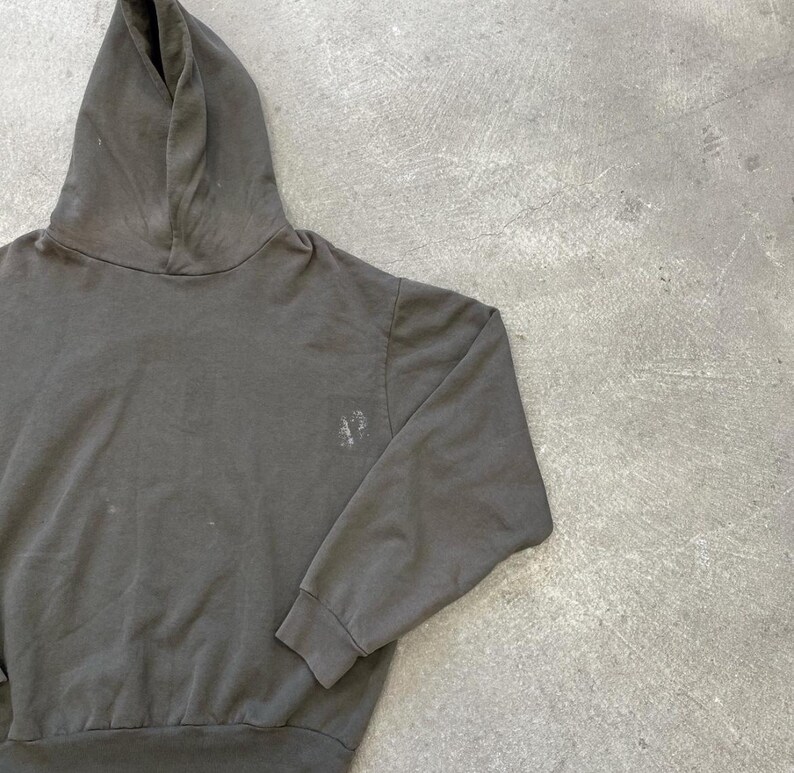 May include: A dark gray hoodie with a drawstring hood and long sleeves. The sweatshirt has a small, faded design on the left chest area. The fabric appears to be a soft, worn material, suggesting comfort and casual wear.