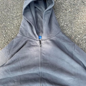 May include: A gray zip-up hoodie with a large hood. The hoodie has a full-length zipper and a small tag at the neckline. The fabric appears to be a soft, washed material. The hoodie is a solid color with no visible graphics or patterns.