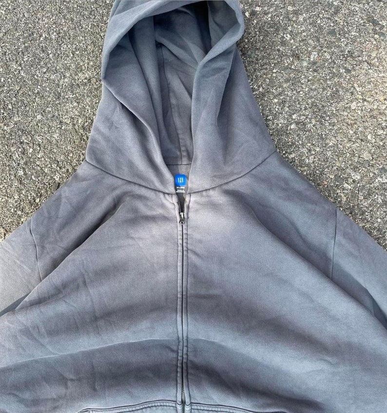 May include: A gray zip-up hoodie with a large hood. The hoodie has a full-length zipper and a small tag at the neckline. The fabric appears to be a soft, washed material. The hoodie is a solid color with no visible graphics or patterns.