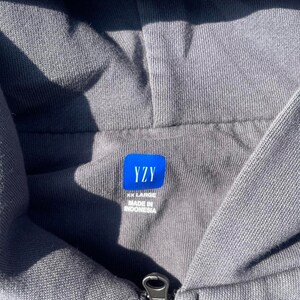 May include: Close-up of a gray hooded sweatshirt with a zipper. A blue label with the white text "YZY" is visible, along with "XX LARGE" and "MADE IN INDONESIA". The fabric has a textured appearance.