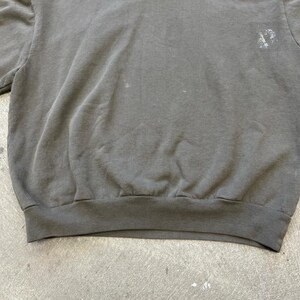 May include: A faded charcoal gray sweatshirt with a crew neck and ribbed hem. The fabric shows signs of wear and tear, with some light-colored spots. The sweatshirt has a simple, classic design.
