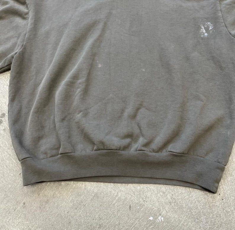 May include: A faded charcoal gray sweatshirt with a crew neck and ribbed hem. The fabric shows signs of wear and tear, with some light-colored spots. The sweatshirt has a simple, classic design.