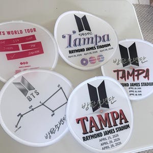 May include: Five round white hand fans featuring BTS concert graphics. Designs include the BTS logo, the text "BTS WORLD TOUR," "TAMPA," "RAYMOND JAMES STADIUM," and "APRIL 25, 2026, APRIL 26, 2026, APRIL 28, 2026," along with member names and constellations.