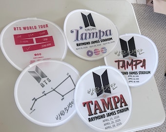 May include: Five round white hand fans featuring BTS concert graphics. Designs include the BTS logo, the text "BTS WORLD TOUR," "TAMPA," "RAYMOND JAMES STADIUM," and "APRIL 25, 2026, APRIL 26, 2026, APRIL 28, 2026," along with member names and constellations.
