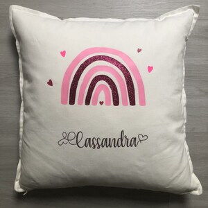 Op de afbeelding: Een wit kussen met een roze en bordeauxrode glitter regenboog en de naam "Cassandra" in cursief geschreven met een hart aan elke kant.