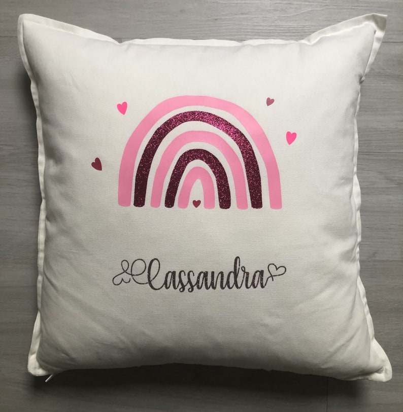 Op de afbeelding: Een wit kussen met een roze en bordeauxrode glitter regenboog en de naam "Cassandra" in cursief geschreven met een hart aan elke kant.