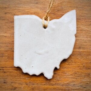 May include: White ceramic ornament shaped like the state of Ohio, with a hole at the top for hanging. The ornament is on a wooden surface.