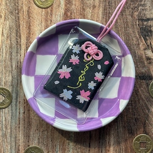 May include: A black fabric charm with pink and white embroidered flowers and the word "Love" in cursive. The charm is attached to a pink cord and is displayed on a purple and white checkered plate.