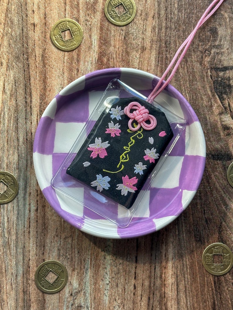 May include: A black fabric charm with pink and white embroidered flowers and the word "Love" in cursive. The charm is attached to a pink cord and is displayed on a purple and white checkered plate.