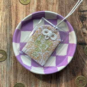 May include: A white and gold Japanese charm with a white tassel, sitting on a purple and white checkered dish. The charm has a gold design and the text "福" in gold.