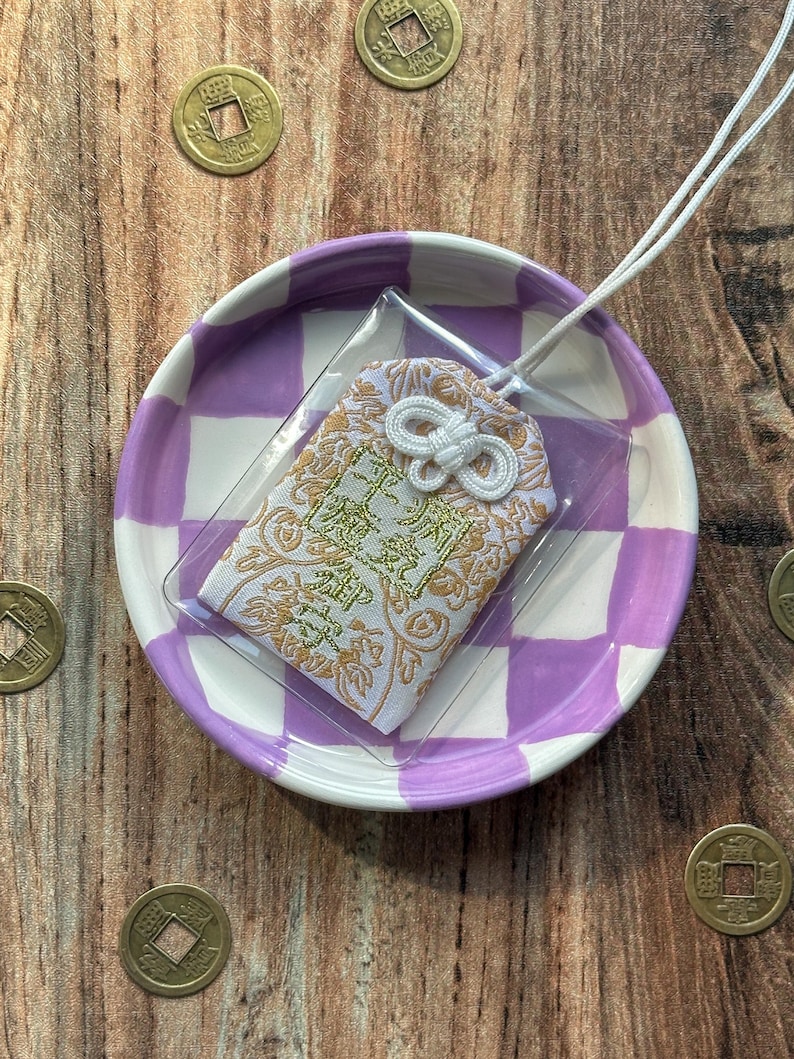 May include: A white and gold Japanese charm with a white tassel, sitting on a purple and white checkered dish. The charm has a gold design and the text "福" in gold.