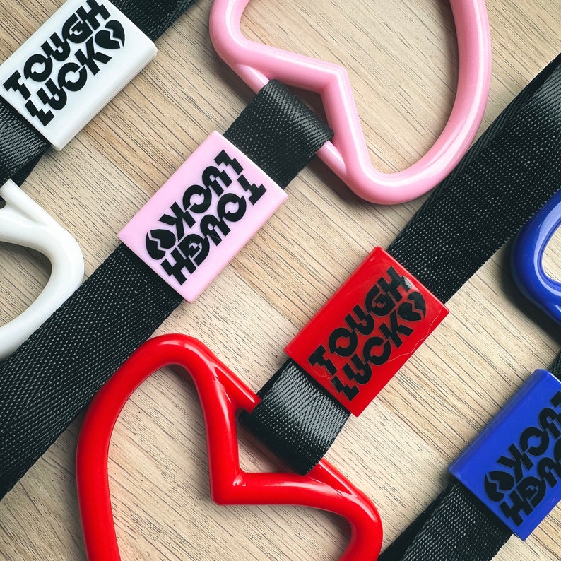 May include: Four car seat headrest hooks with black straps and pink, red, and blue heart-shaped charms. Each charm has the text "Tough Luck" printed on it.