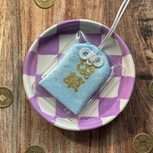 May include: A light blue Japanese charm with gold lettering and a white tassel, displayed on a purple and white checkered plate.