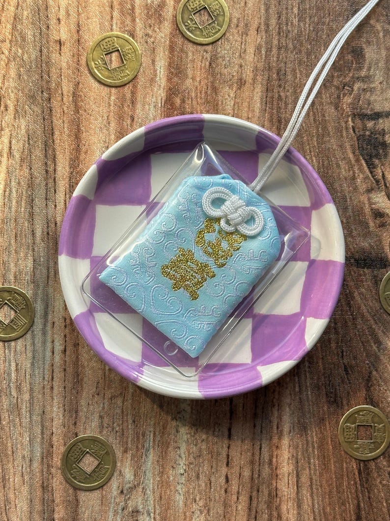May include: A light blue Japanese charm with gold lettering and a white tassel, displayed on a purple and white checkered plate.
