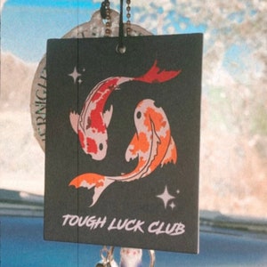 May include: Black car air freshener featuring two orange and white koi fish with the text "Tough Luck Club".
