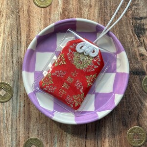 May include: A red and gold Japanese charm with a white string, displayed on a purple and white checkered plate. The charm is in a clear plastic bag.