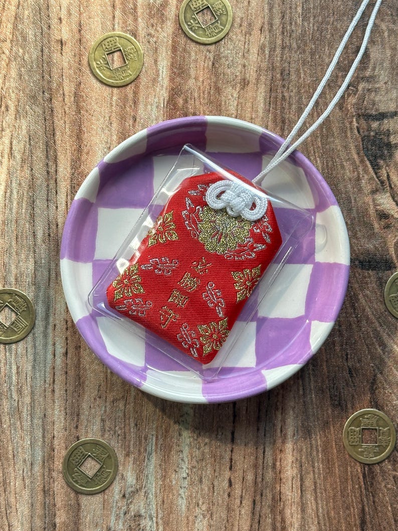 May include: A red and gold Japanese charm with a white string, displayed on a purple and white checkered plate. The charm is in a clear plastic bag.