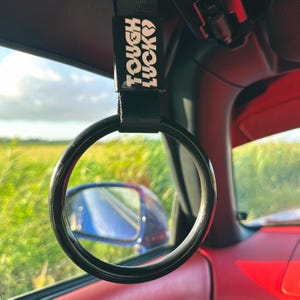 May include: Black car interior with a black and white "Tough Luck" strap hanging from the ceiling. The strap has a black ring attached to it.