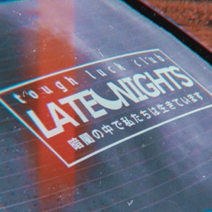 May include: A black and white sticker with the text "tough luck club LATE NIGHTS" and Japanese text that reads "暗闇の中で私たちは生きています" which translates to "We live in the darkness".