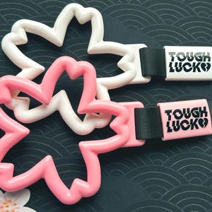 Może przedstawiać: Dwa plastikowe breloczki w kształcie kwiatu w kolorze r&oacute;żowym i białym z czarnymi paskami. Każdy pasek ma białą lub r&oacute;żową prostokątną etykietę z tekstem "TOUGH LUCK" w kolorze czarnym.