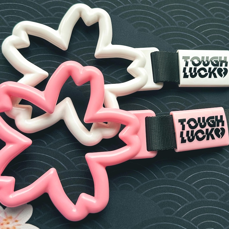 Może przedstawiać: Dwa plastikowe breloczki w kształcie kwiatu w kolorze r&oacute;żowym i białym z czarnymi paskami. Każdy pasek ma białą lub r&oacute;żową prostokątną etykietę z tekstem "TOUGH LUCK" w kolorze czarnym.