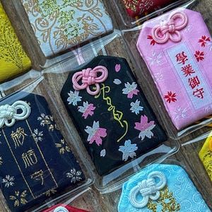 May include: Assortment of colorful Japanese good luck charms, or "omamori", in various fabrics and designs. Each charm features a different inscription in Japanese characters, including "学業 御 守" (Gakugyo Omamori) for academic success, "厄除け" (Yakuyoke) for protection from misfortune, and "商売繁盛" (Shobai Hanjo) for business prosperity.
