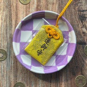 May include: A yellow fabric charm with gold embroidery and a yellow string tied in a knot. The charm is on a purple and white checkered plate with gold coins scattered around it.