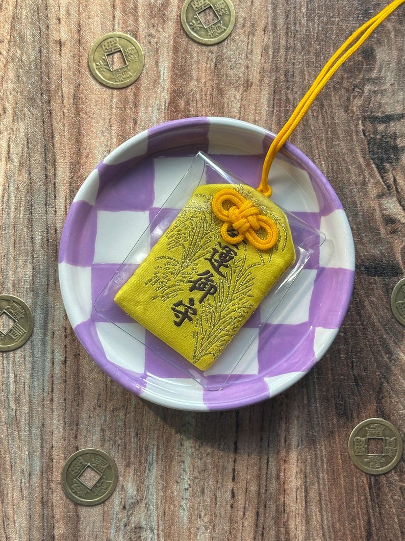 May include: A yellow fabric charm with gold embroidery and a yellow string tied in a knot. The charm is on a purple and white checkered plate with gold coins scattered around it.