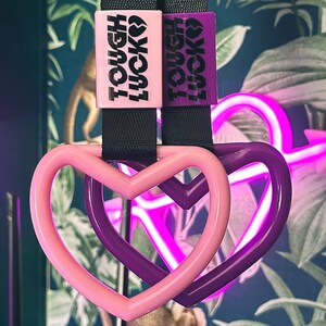 May include: Two purple and pink heart-shaped keychains with black straps. The keychains have the text "Tough Lucky" printed on them.