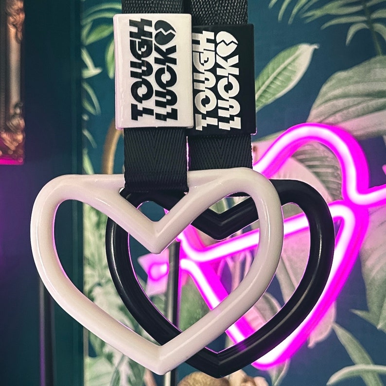 May include: Two black keychains with white heart-shaped charms. The keychains have the text "Tough Luck?" printed on them. The charms are attached to a white heart-shaped charm with a black heart-shaped charm inside.