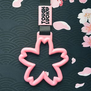 Może przedstawiać: R&oacute;żowy plastikowy brelok do samochodu w kształcie kwiatu wiśni z czarnym paskiem z napisem "Tough Luck".