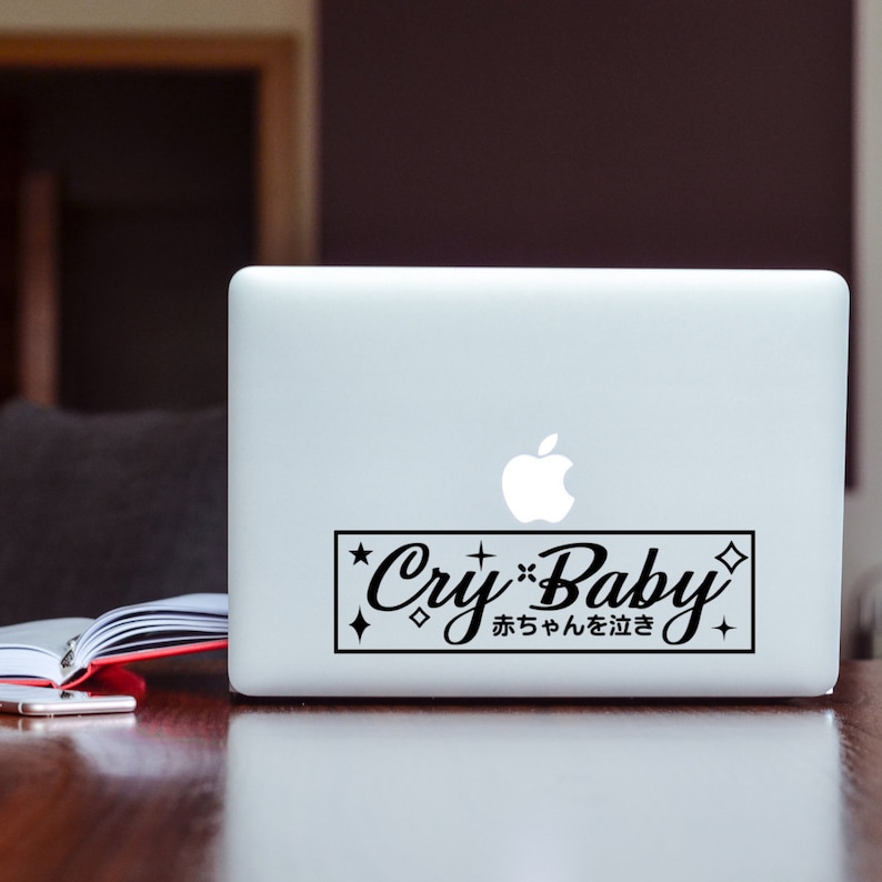 May include: A black and white sticker on a laptop. The sticker has the words "Cry Baby" in a stylized font, with Japanese characters below. The design is framed by a black rectangle with star and diamond accents.