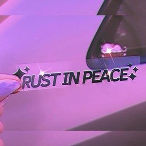 May include: A clear sticker with the text "RUST IN PEACE" in black lettering with two small black diamond shapes on either side of the text. The sticker is being held by a hand against a purple background.